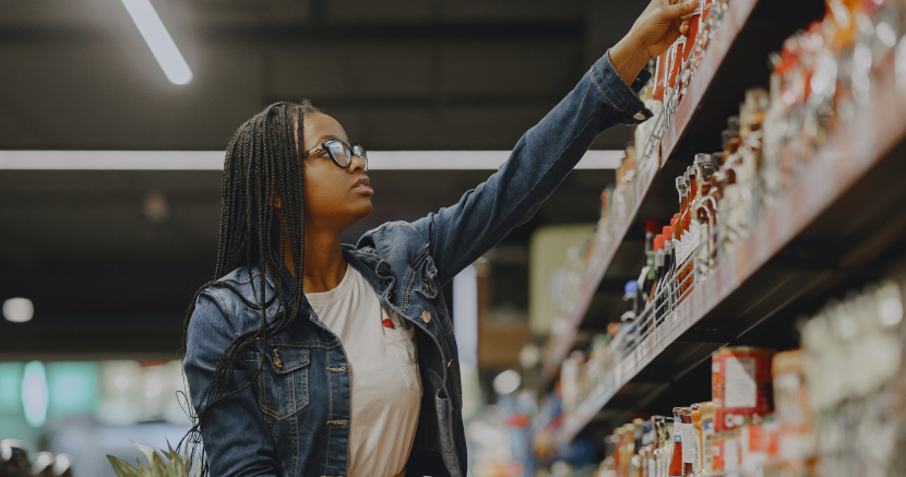 A young woman with braids and glasses reaches for a box on a grocery store shelf.