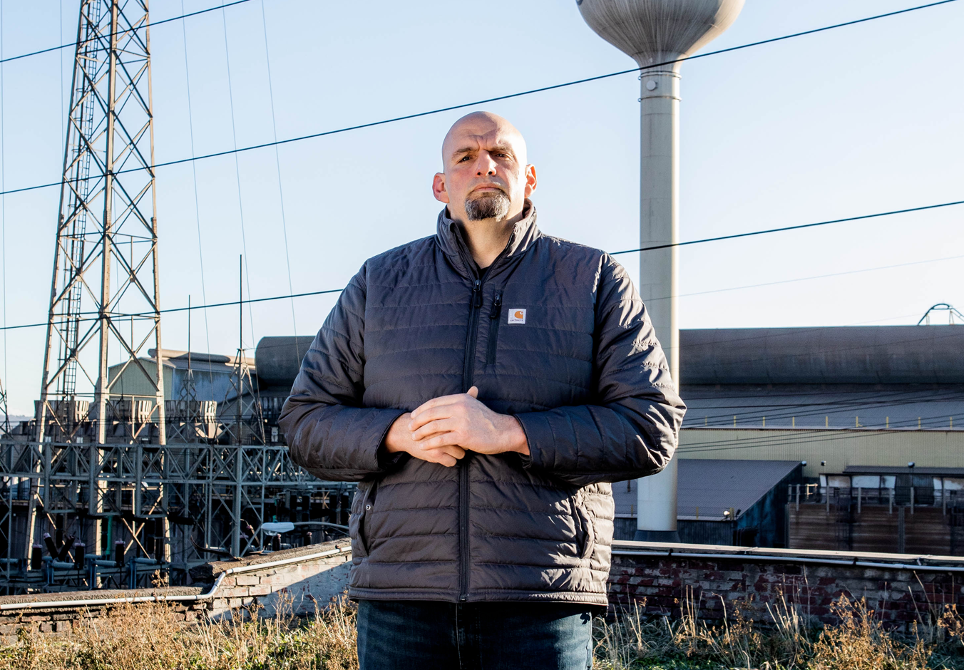John Fetterman stands with hands folded in front of a water tower and electrical lines. 