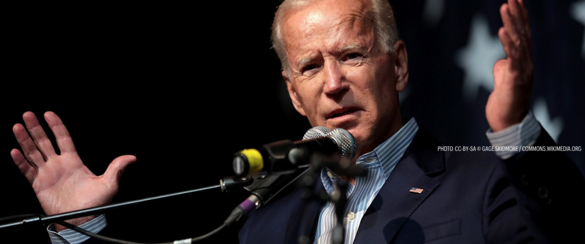 President Biden stands before a microphone with his hands outstretched.