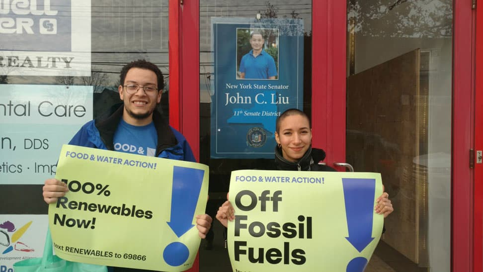 Two young volunteers stand with signs that read "100% Renewables now!" and "Off fossil fuels." They stand in front of a building with a poster that reads "New York State Senator John C. Liu 11th Senate District" in the window.